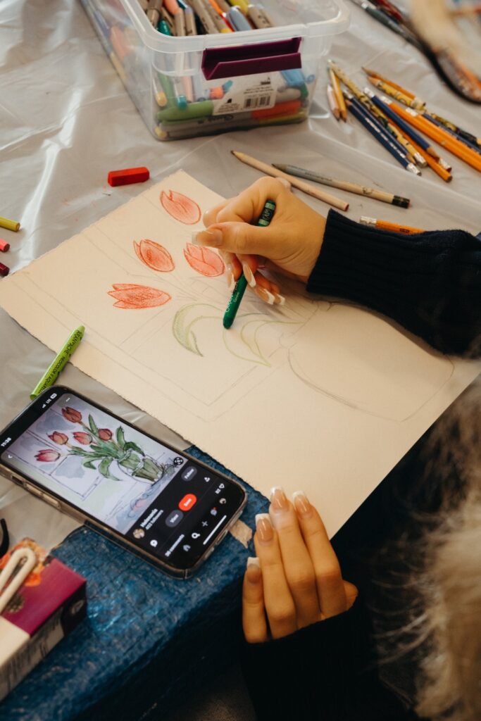 Hands hold crayons and sketch flowers onto a white paper. A phone sits next to the paper with a reference image of the same flowers.