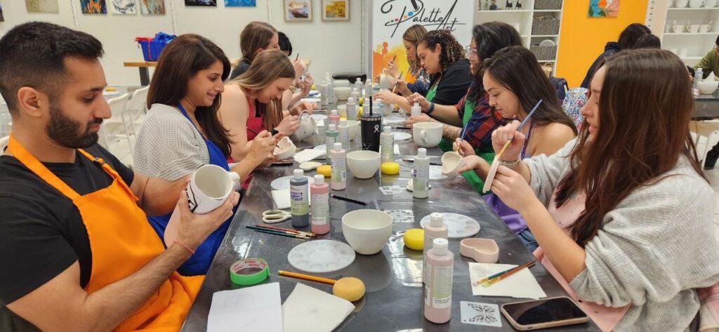 A long table stretches the length of the room. People seated on either side paint various pieces of pottery. The table is covered with paints, brushes, and cups of water.